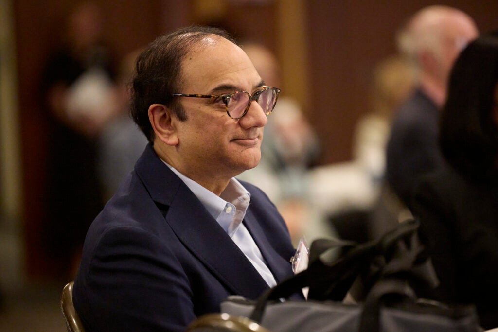 A man in a dark suit looks attentively through horn-rimmed glasses. 