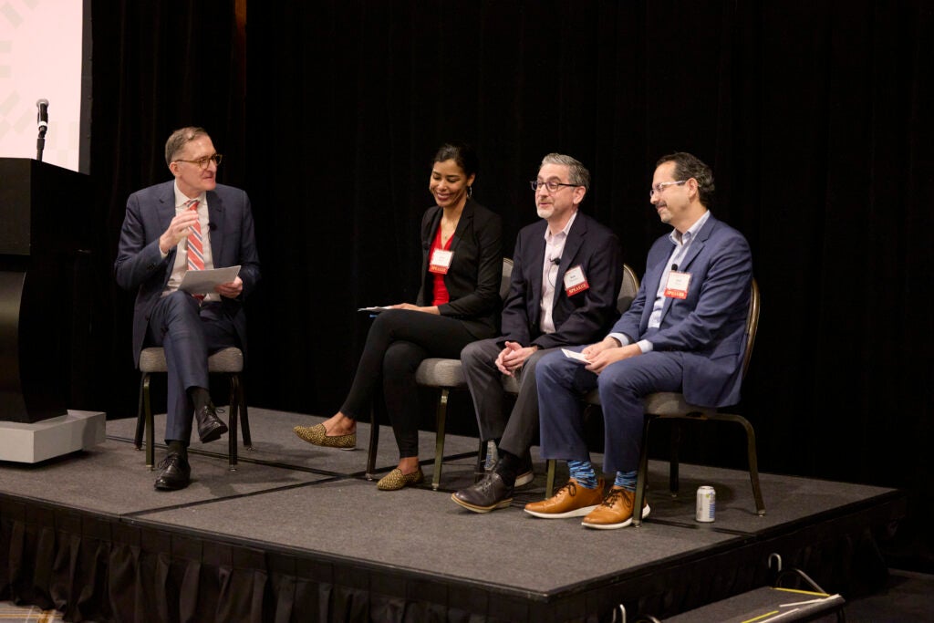 The Dean, seated separately to the left of the image, asks a question while gesturing with his right hand. The speakers are seated in a row, and their faces show reactions of mirth and thoughtfulness. 