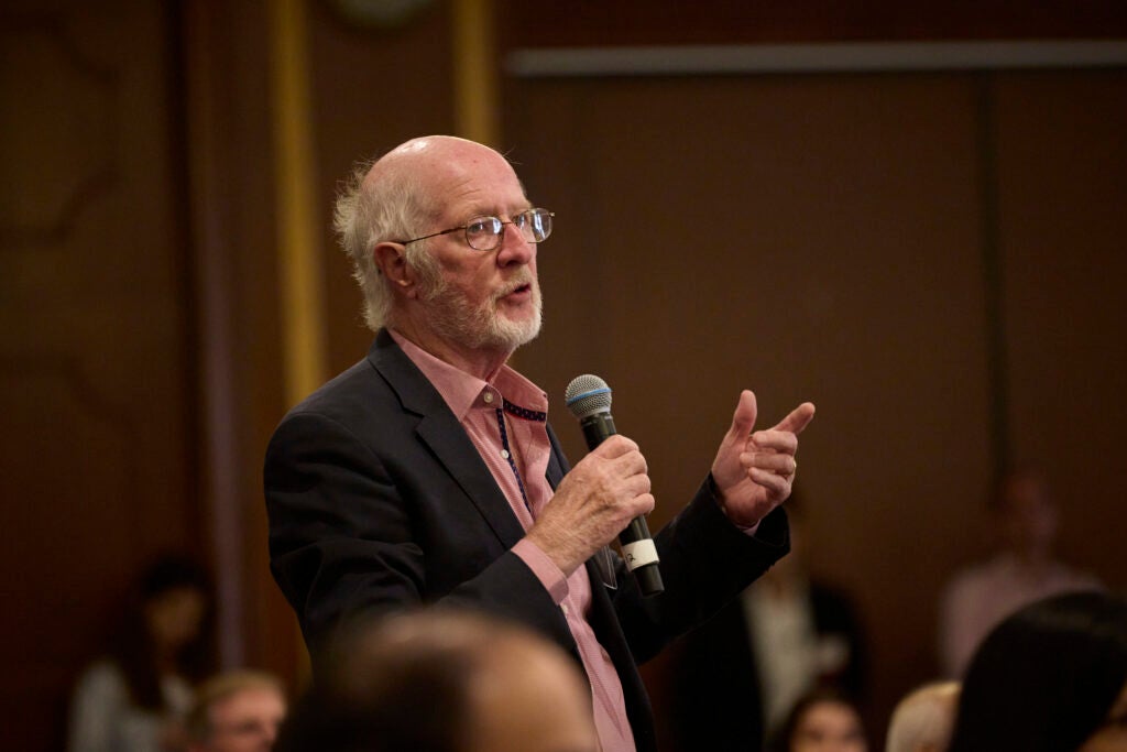 A man wearing glasses, a black suit, and a red shirt stands to ask a question. 