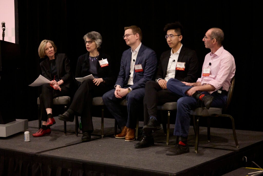 The moderator and speakers are all seated in a row. Susanna Baruch,  second from left, is speaking, and the others are focused on her. 