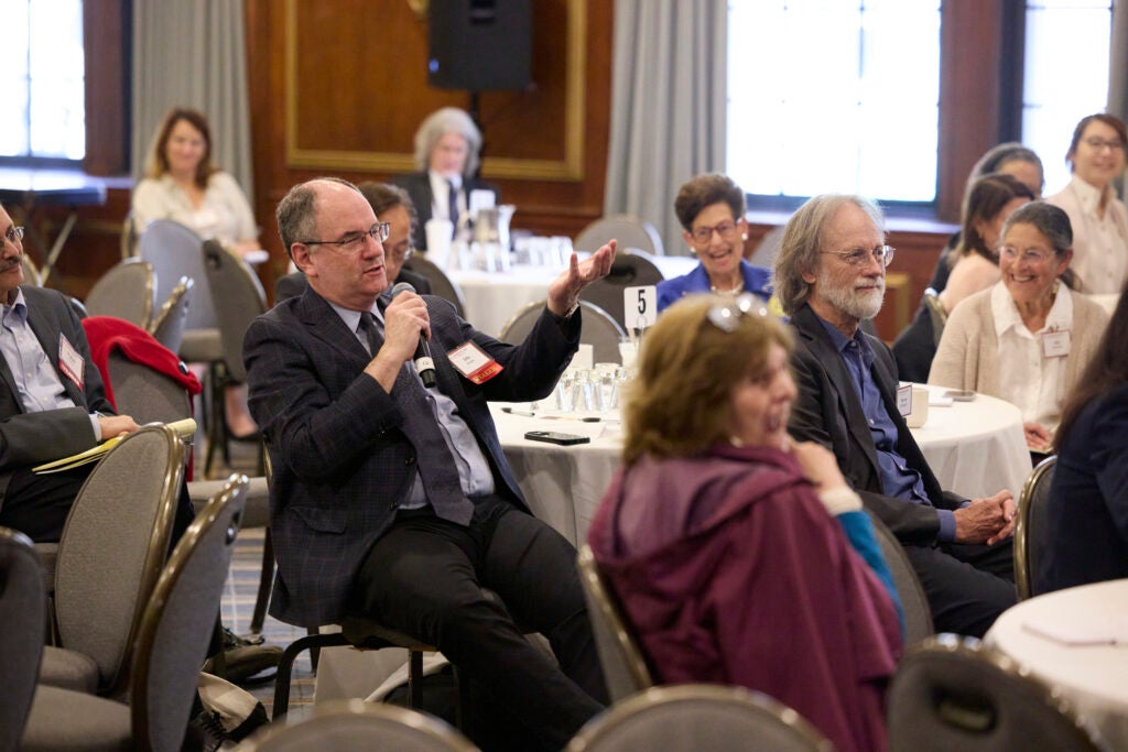 A men wearing glasses and a dark suit asks a questions from a table. Several other audience members in the image are smiling and laughing while he speaks. 
