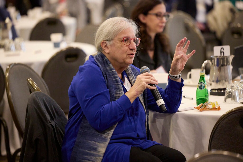 A woman wearing red-rimmed glasses and a bright blue top asks a question from her table. 