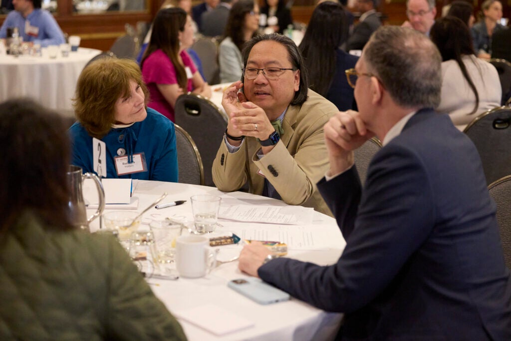 A man in a khaki suit speaks emphatically while those on either side turn to engage. 
