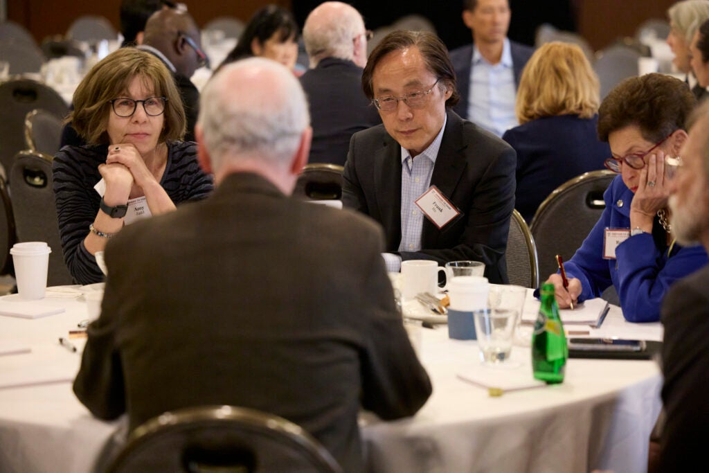 Two attendees watch a third, who we see from the back. On the right, another is taking notes with a crimson pen. 