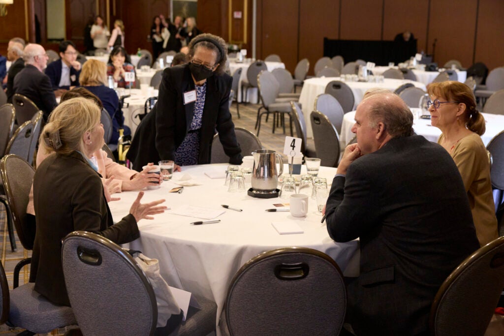 One woman leans on a table, half-standing, while listening to another speak with her hands spread in gesture. The rest of the table looks on. 