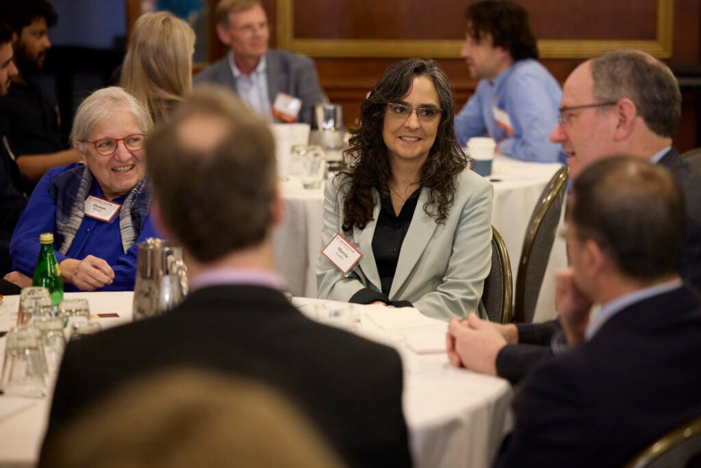 Guests at a table smile while watching a tablemate speak in the right of frame. 