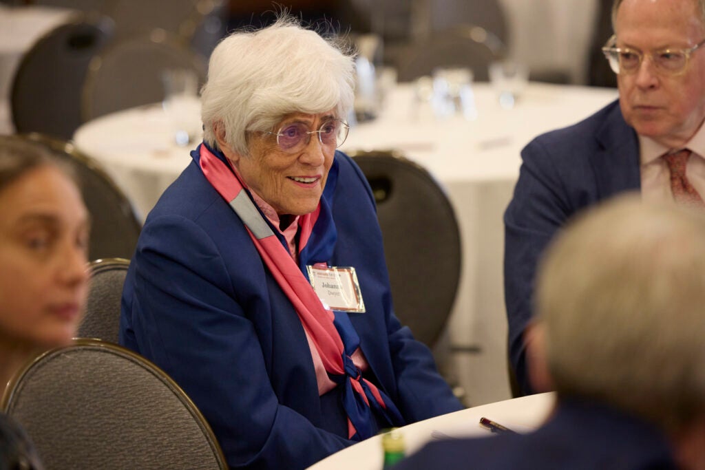 A woman in a bright blue jacket, pink patterned scarf, and glasses smiles at her table