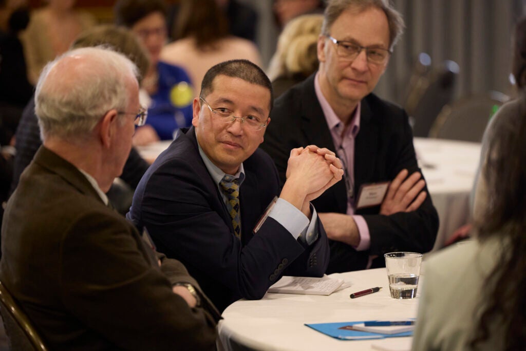 Two men in suits and glasses turn to listen to a third, who we see from the back. 