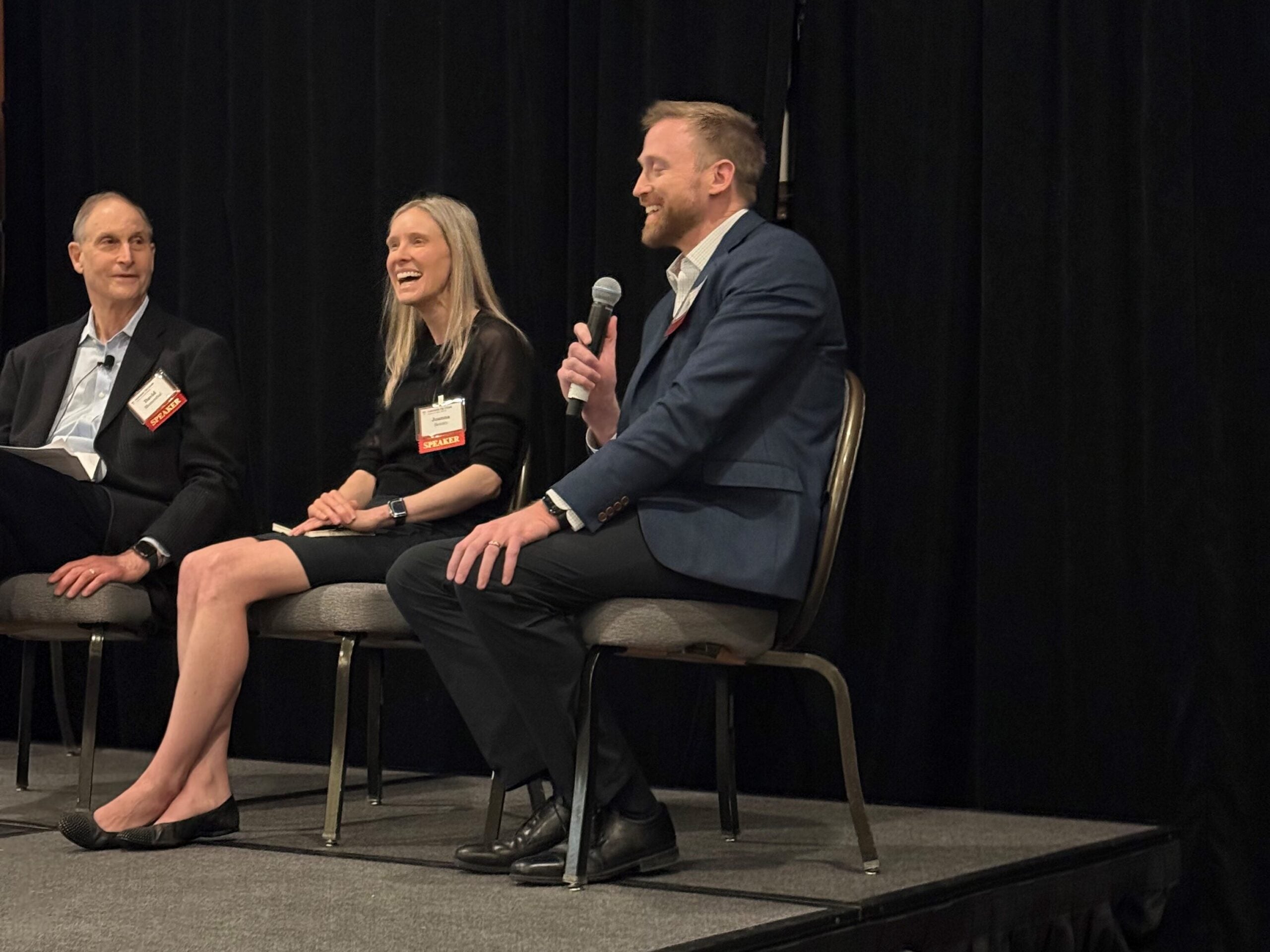 All three are seated. In the foreground, David Bates speaks into the mic with a smile. Joanna Beasley laughs at his right, and David Blumenthal, in the back left, also looks amused.  