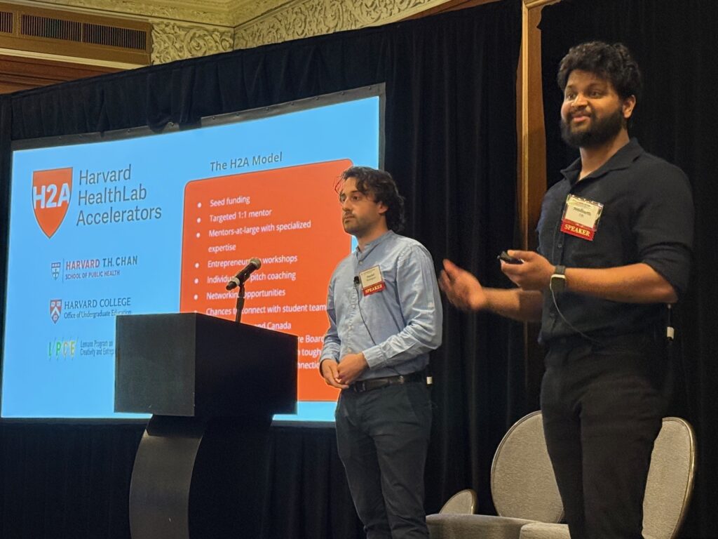 Two students are on stage with a  slide reading "Harvard HealthLab Accelerators" behind them. One, with collar-length hair and a blue shirt, is at the podium, and the other, with a full beard and a black shirt rolled up to the elbows, gestures while speaking in the foreground. 