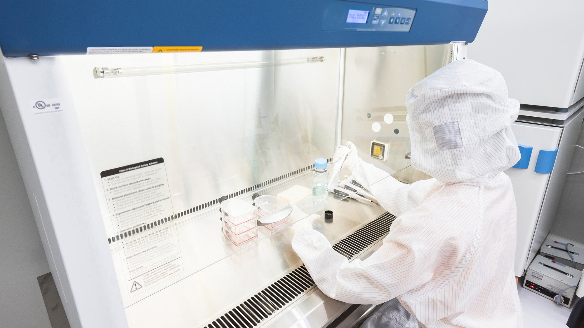 A scientist in sterile coverall gown pipetting medium or reagents for cell culture experiment in biological safety cabinet.