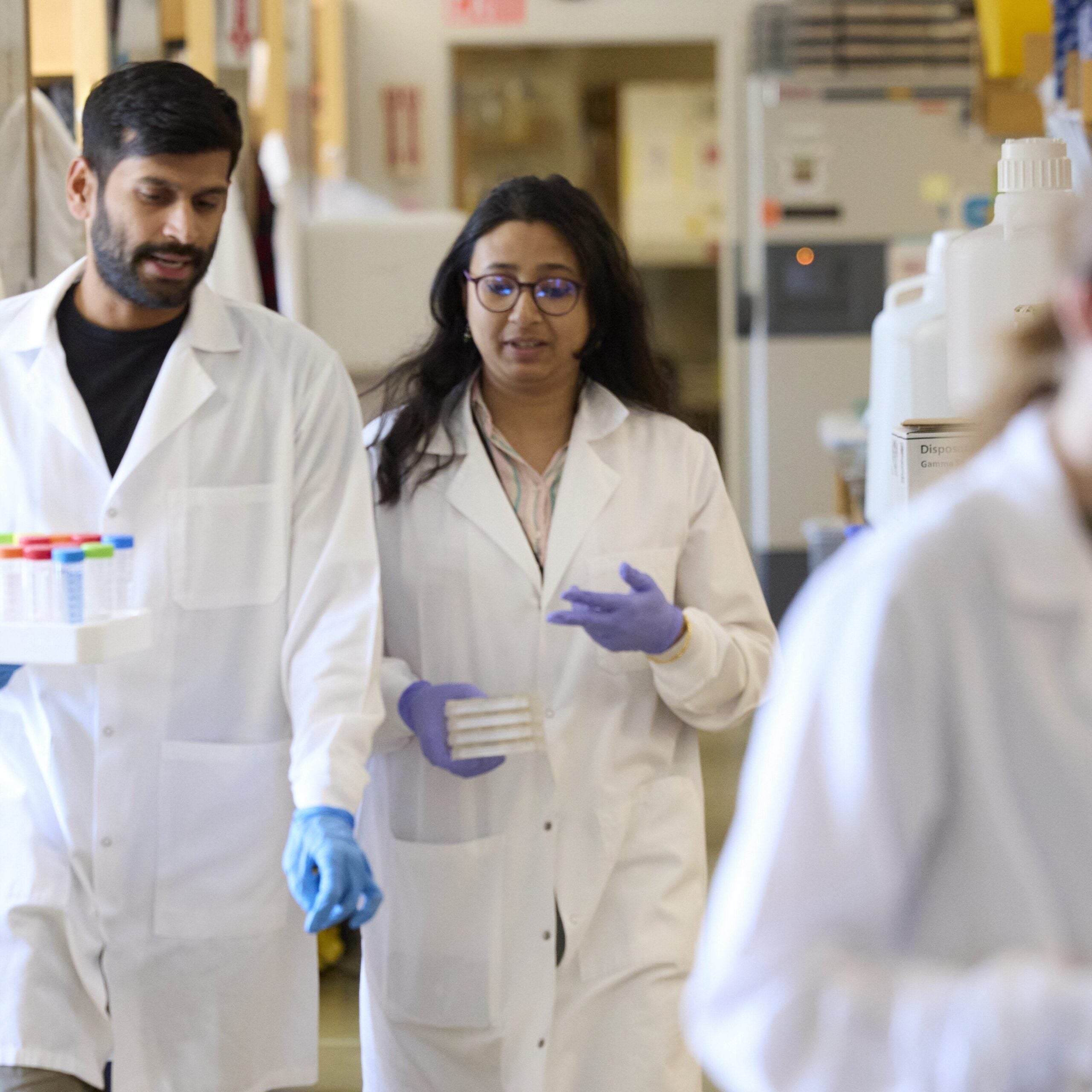 Two lab colleagues converse while walking down the aisle holding small pieces of equipment. In the foreground, another colleague is busy at a bench.
