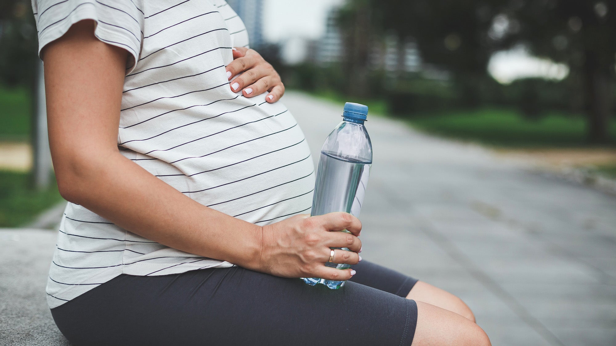 A pregnant woman sits on a bench in the park in the summer and holds a bottle of water in her hand. Her face is not visible.
