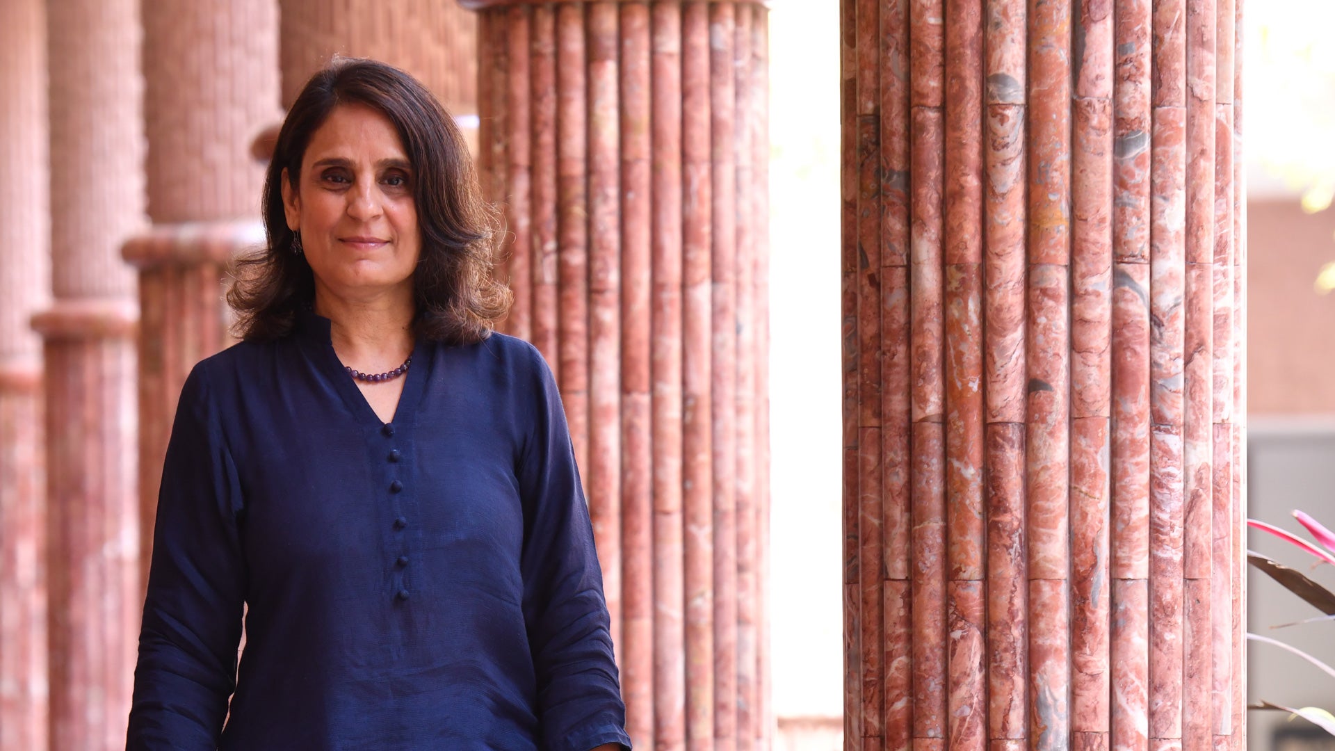 Salimah Walani walks through a columned courtyard, looking at the camera.