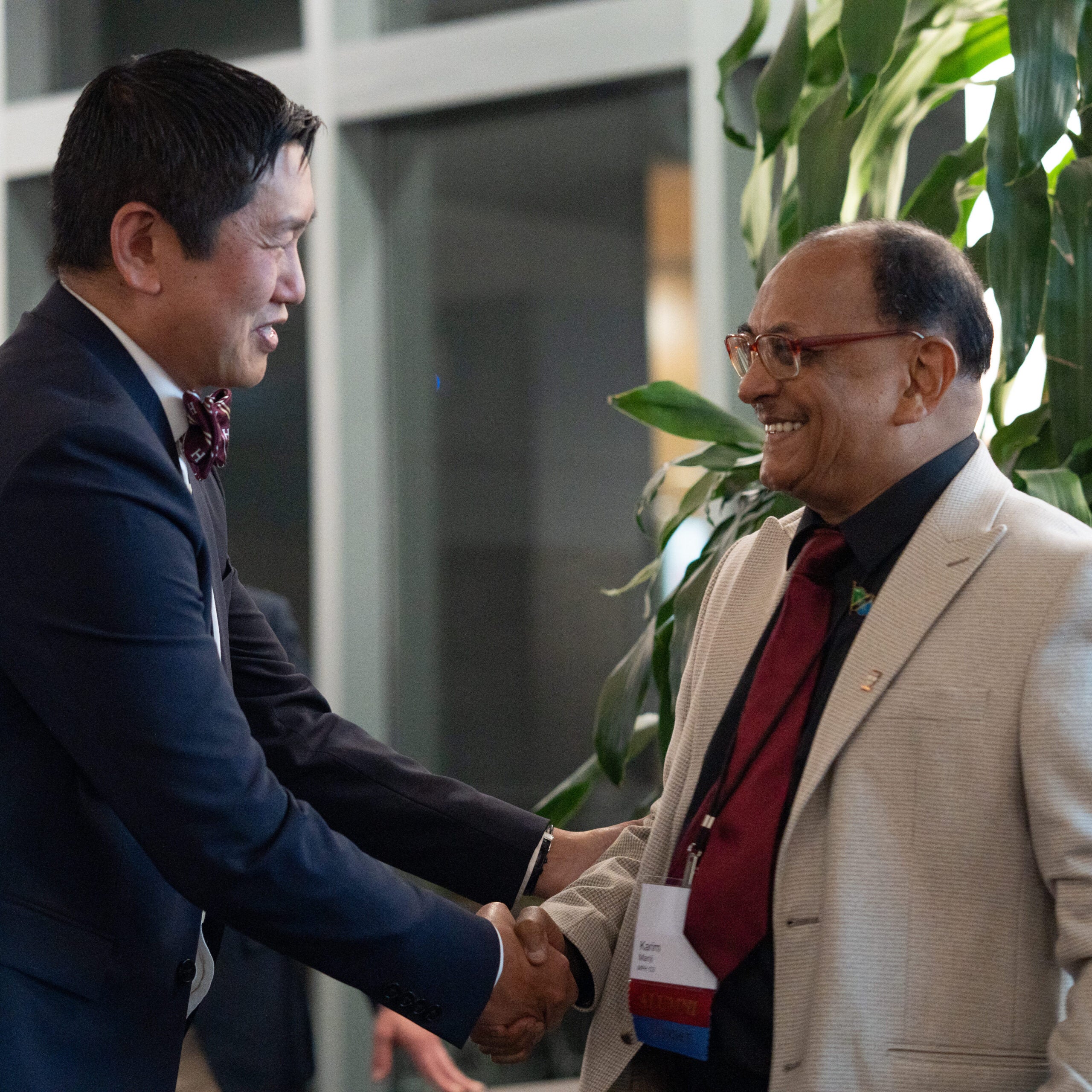 Both smiling, a man in a blue suit and Harvard bowtie shakes hands with a man in a cream suit and red tie.