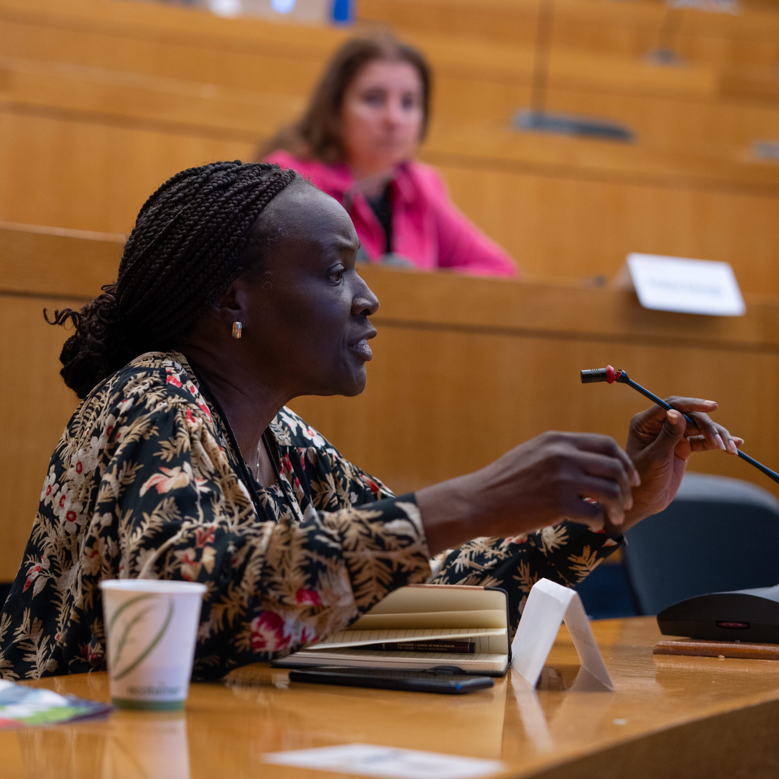 A woman with thin braids pulled into a low bun speaks earnestly into the microphone at hear seat in a lecture hall.