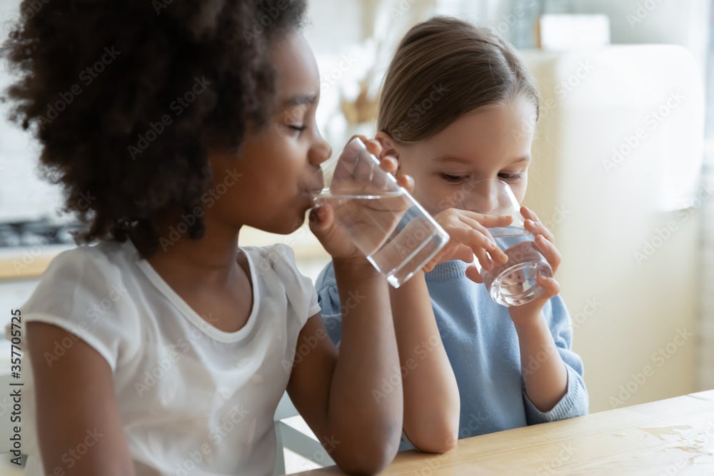 Two multi racial little girls sit at table in kitchen drinking glasses of water.