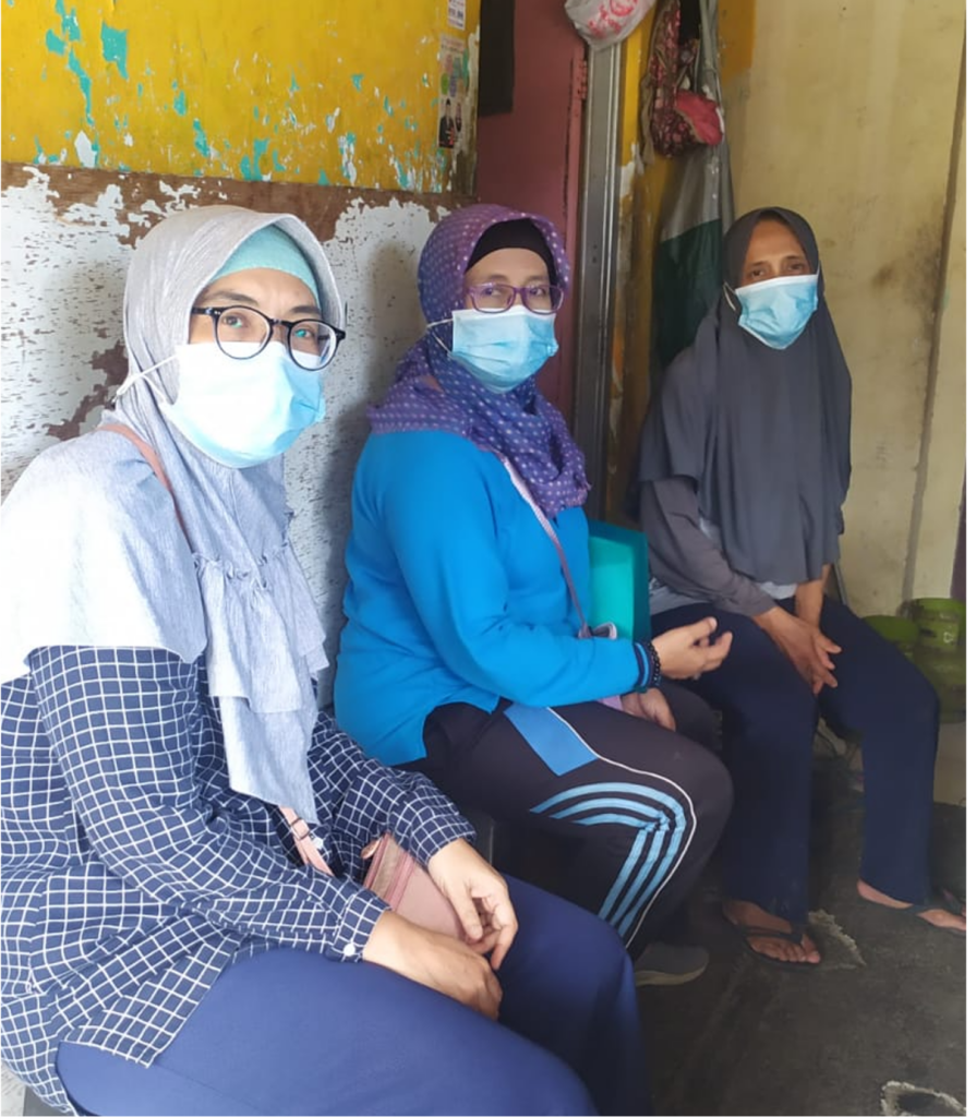Three women wearing head coverings and Covid-19 masks sit on a bench, facing the photo-taker.