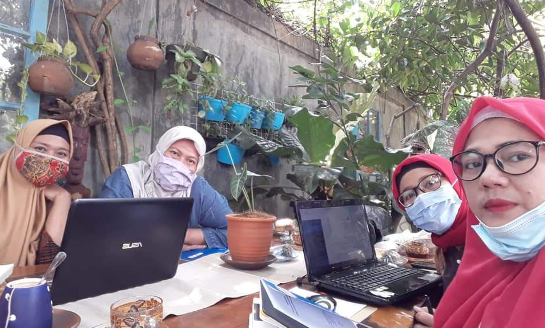 Four women sit around a table with laptops. They are wearing head coverings and Covid-19 masks.