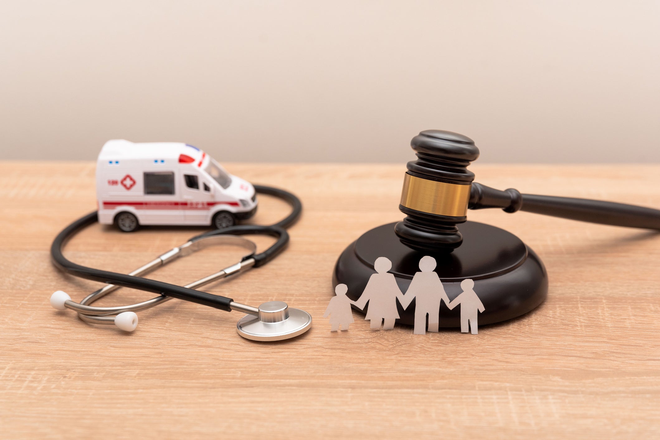 On a wooden table, a stethoscope wraps around a toy ambulance and paper figures of a family stand in front of a gavel.