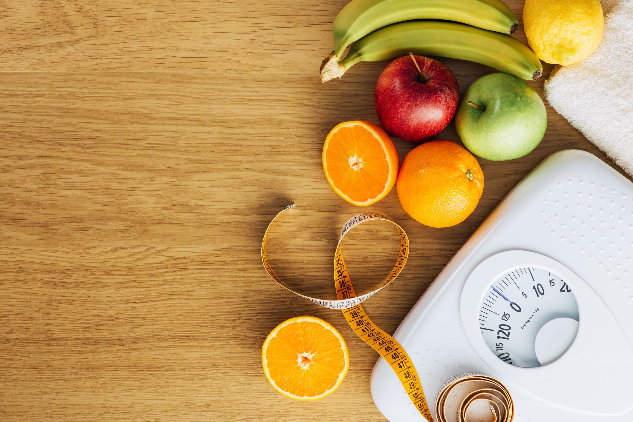 A while scale sits on a wood table surrounded by apples, bananas, and oranges, as well as a soft measuring tape.