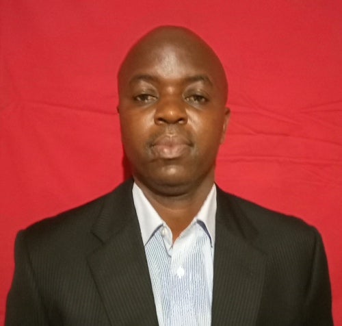 Headshot of Miller Alusa in front of a red background wearing a black tuxedo.