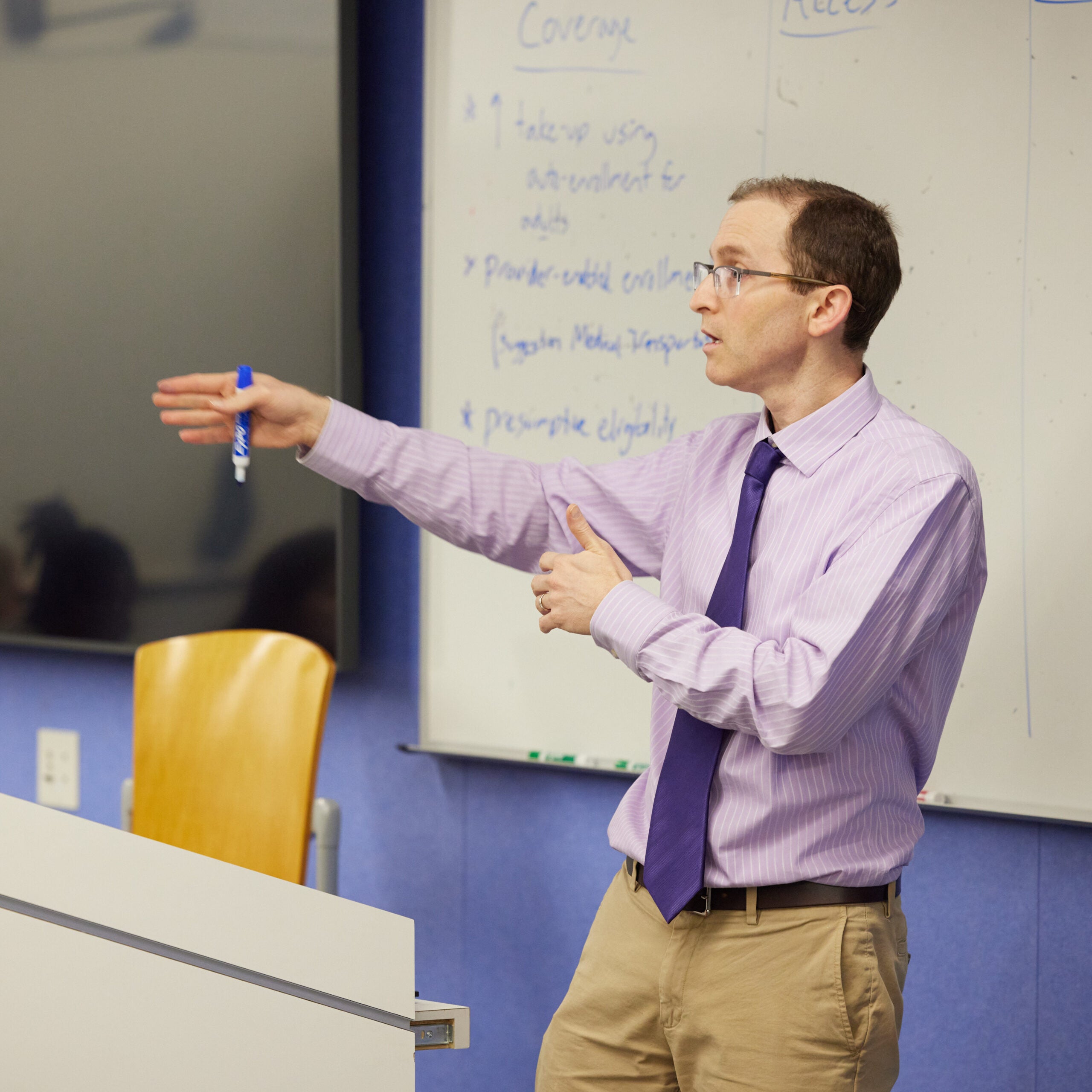 Ben Somers speaks at a podium, holding a dry-erase marker in one outstretched hand. His whiteboard notes are out of focus in the background. 