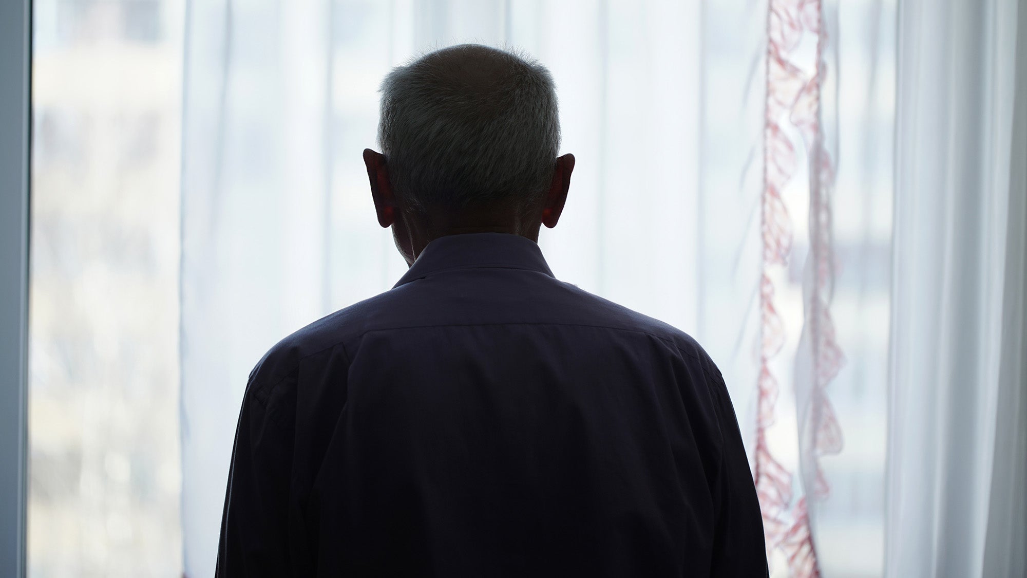 Silhouette of older man looking through window with transparent curtain