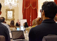 Audience members seated with laptops listen to a speaker in a chandelier-lit room with red curtains, during a leadership course session.