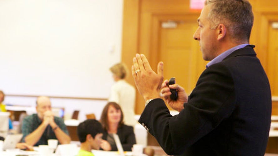 A professor in a suit gives a presentation in a classroom during a sustainability program course, speaking and gesturing with his hands while holding a remote, as several seated attendees listen and take notes in the background.