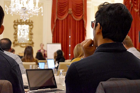 Audience members seated with laptops listen to a speaker in a chandelier-lit room with red curtains, during a leadership course session.