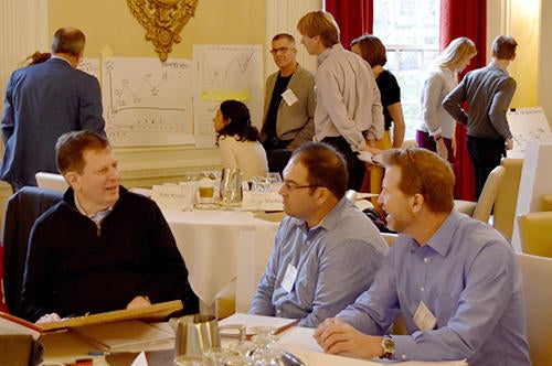 Sustainability Leaders participate in a hands-on exercise engaging in small group discussions at round tables, while others review charts and diagrams displayed on the walls in the background.