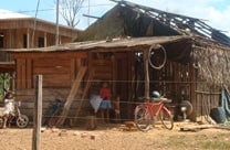 Small wooden house with a partially damaged roof, bicycles in front, and children standing near the entrance on a dirt yard, with another building visible in the background.