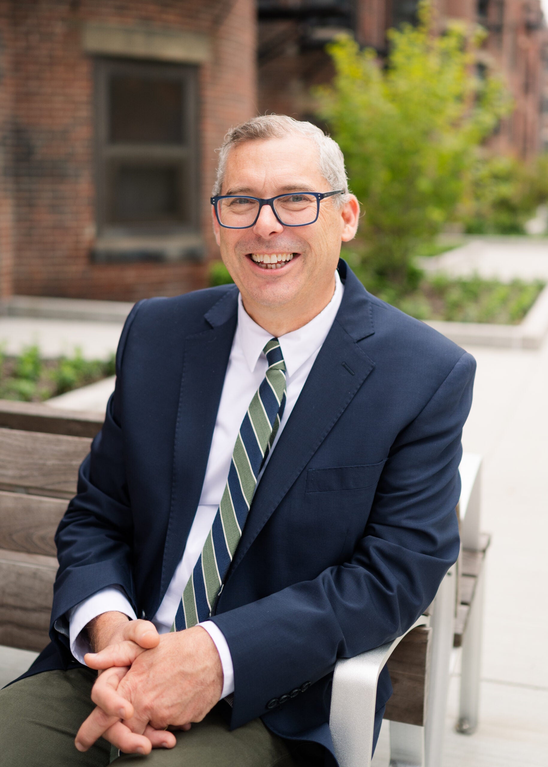 Headshot of a man with white hair and black square glasses, white shirt, striped tie, and blue blazer, sitting in a chair with greenery and a window in the background