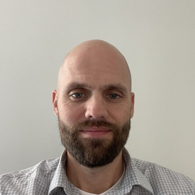 Headshot of a man with a brown beard and bald wearing a white striped shirt with a white background