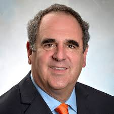 Headshot of a man with gray/brown hair and dark eyebrows with a light blue shirt, orange tie, and dark blue blazer