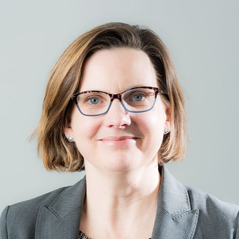 Headshot of a woman with short brown hair, glasses, and gray blazer with a gray background
