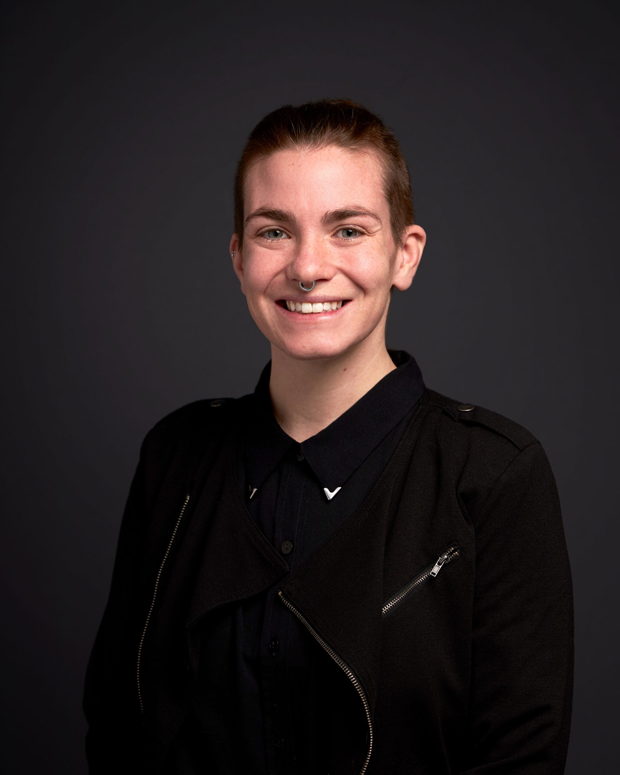 Headshot of person with short brown hair wearing all black with dark gray backdrop