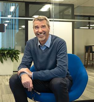 A man with white hair smiling sitting in a chair resting elbows on his legs wearing a blue sweater and sitting in a blue chair
