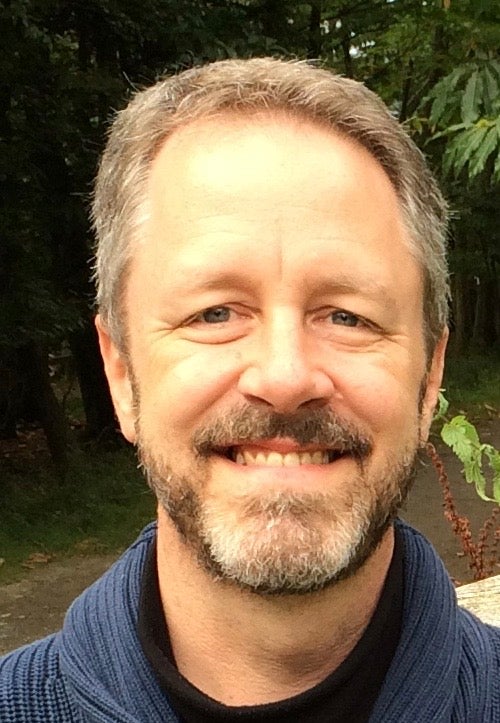 Close up headshot of man with gray hair and gray beard with blue shirt on and greenery in background