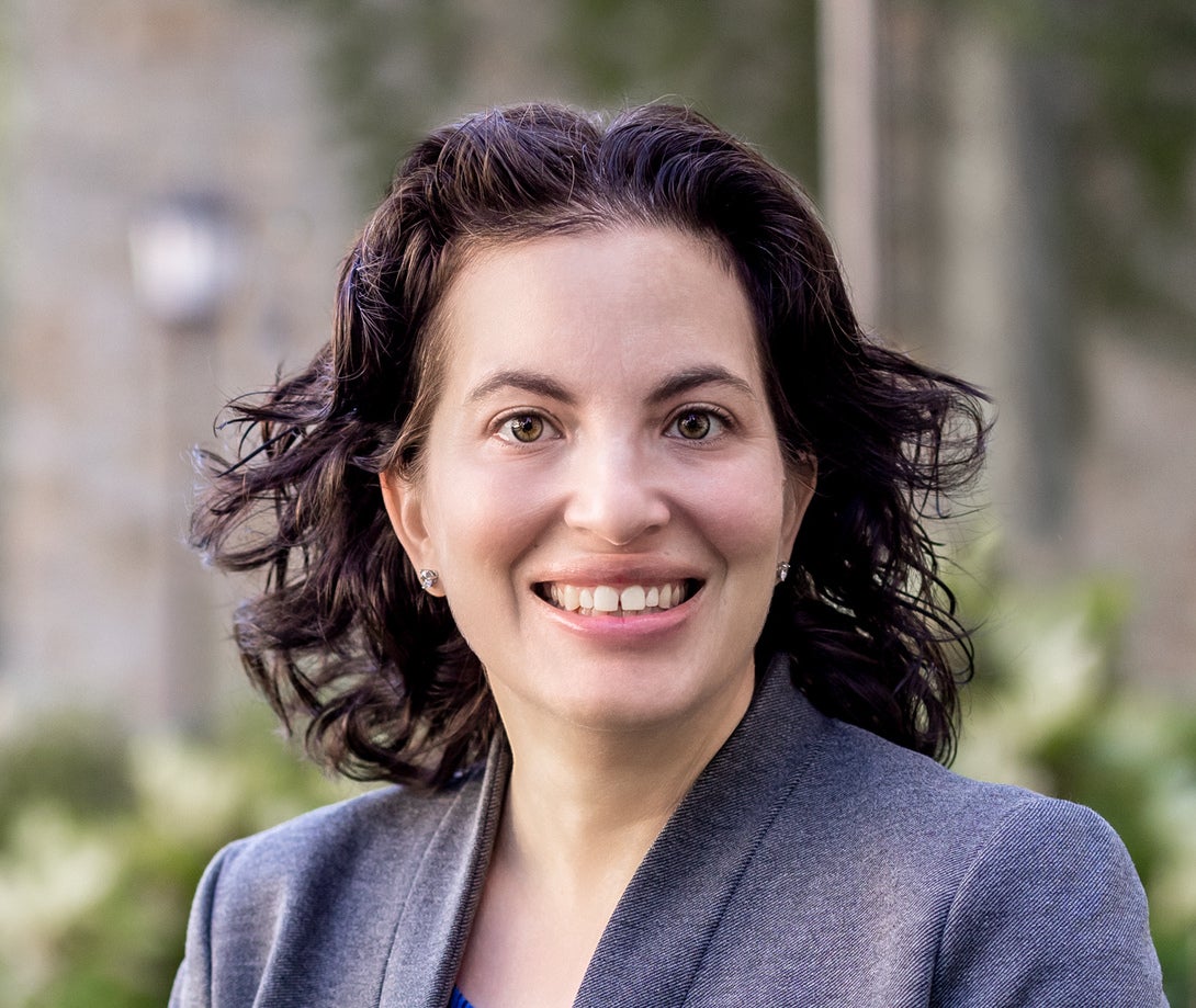 Headshot of woman with short dark curly hair in a gray blazer with greenery in background