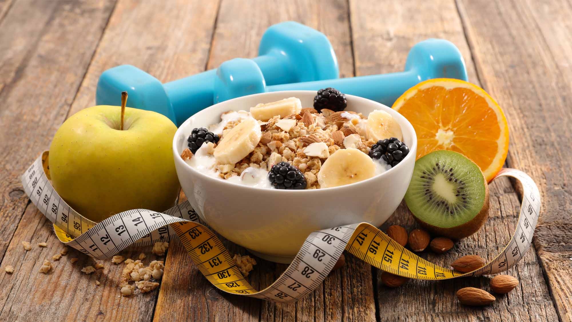 Healthy foods, dumbbells, and a measuring tape are arranged on a table.