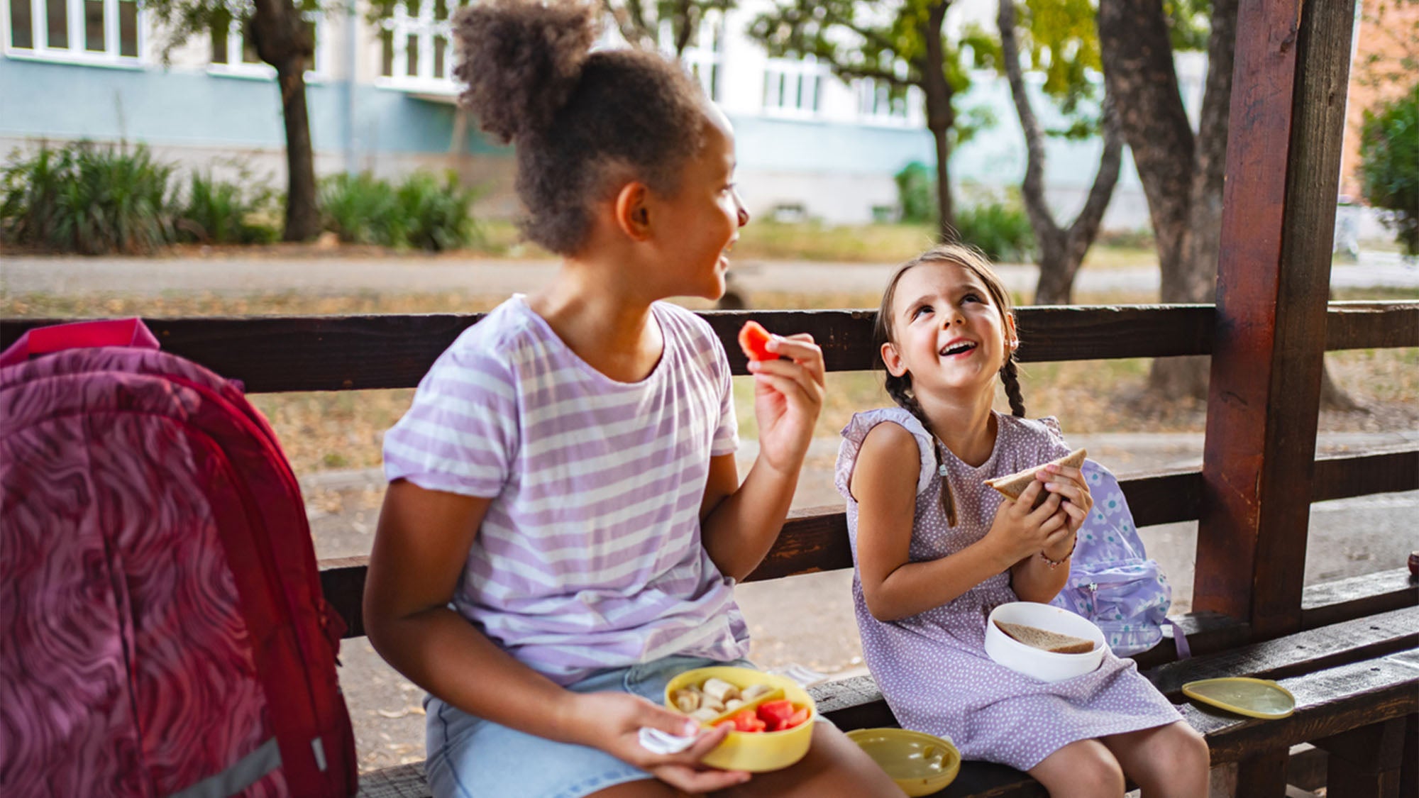 Two schoolchildren eat lunch together on a bench, smiling.