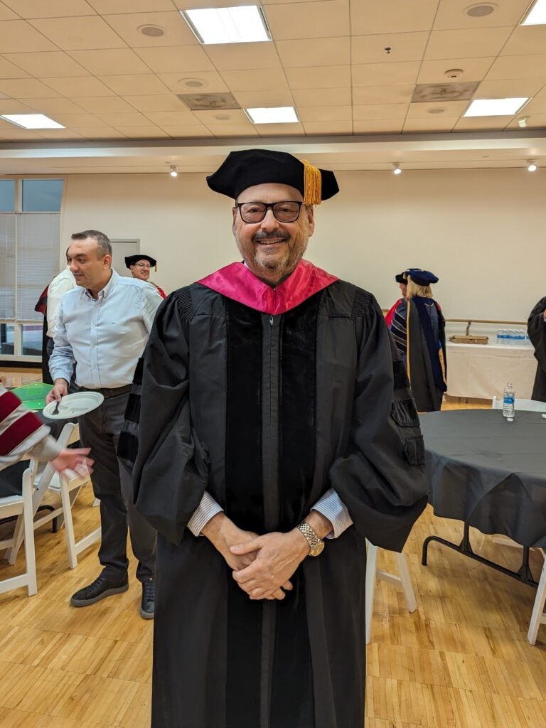 Alan Cohen posing in academic regalia, with the commotion of a commencement behind him.