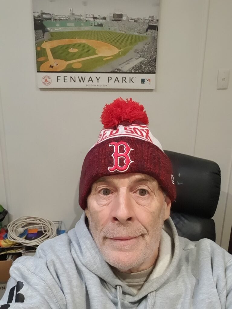 Selfie of Chris Bain wearing a knit Red Sox Cap in front of a poster of Fenway Park.