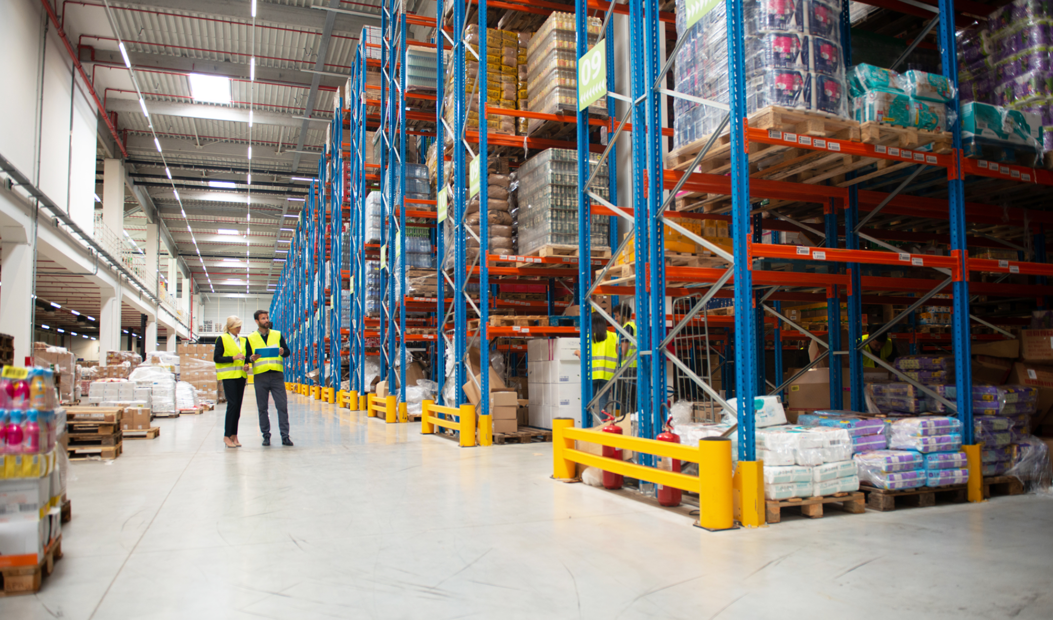 Brightly lit warehouse corridor with tall stacks of boxed goods for distribution, 2 workers standing on the side in yellow safety vests, one man with dark hair, one blonde women