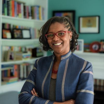 Headshot of woman with short dark curly hair, red glasses, blue blazer, bookshelf in background
