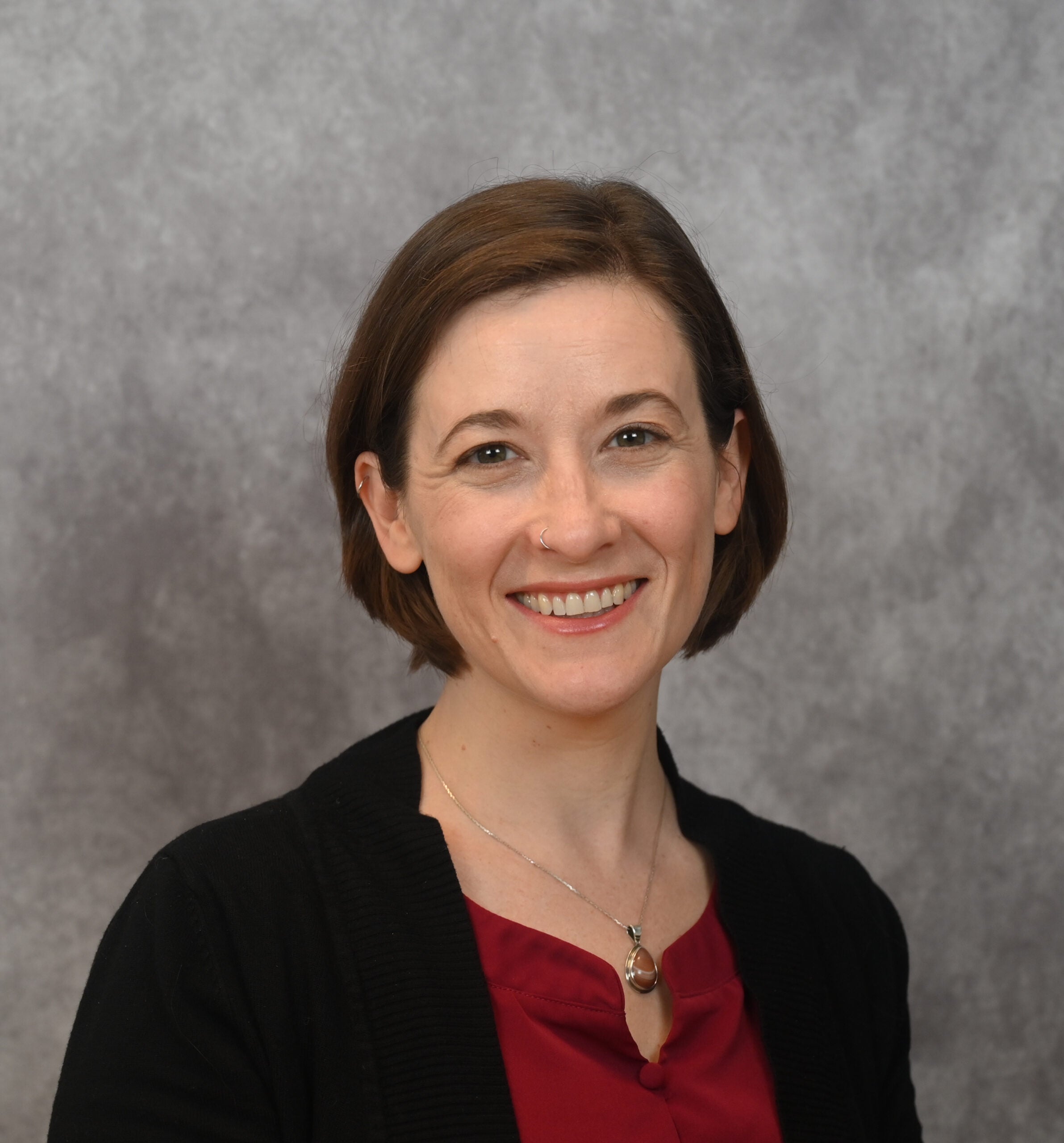 Headshot of woman with short brown hair, red shirt, gray background.