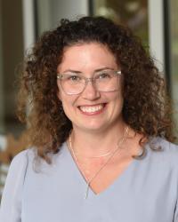 Headshot of woman with brown curly hair, glasses, gray shirt