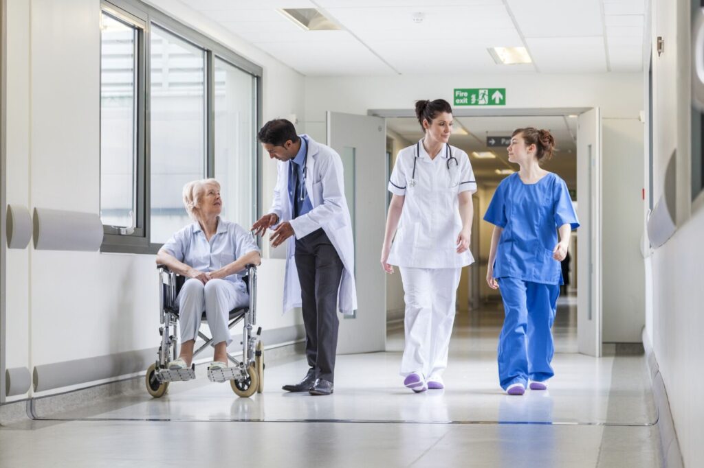 Two doctors in white lab coat and one nurse in blue, walking down a hospital corridor with an elderly person in a wheelchair